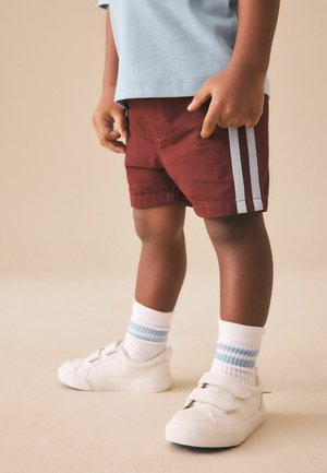 Bambino che indossa pantaloncini color granata con strisce bianche, scarpe da ginnastica bianche con cinturini in velcro e calzini bianchi con strisce blu, in piedi su un pavimento beige.