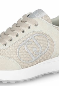 Sneaker beige in materiali scamosciati e mesh, con un logo argento, lacci grigio chiaro e una suola bianca testurizzata. Design con punta rotonda.