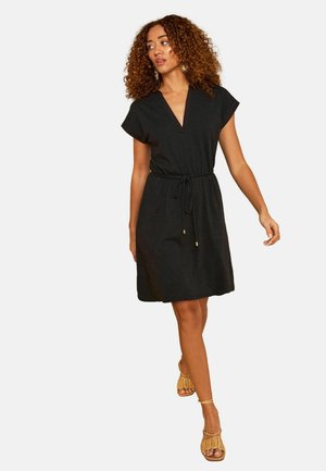 Frau mit lockigem Haar, die ein schwarzes Wickelkleid mit kurzen Ärmeln und Taillengürtel sowie beige, gewebte Sandaletten mit Absatz trägt und vor einem weißen Hintergrund läuft.