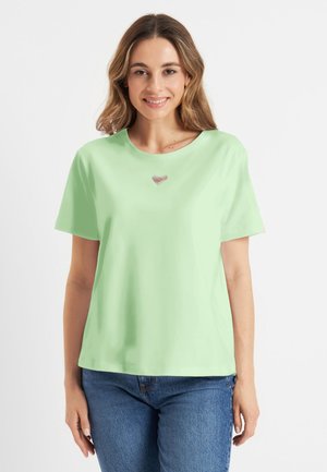 Vrouw met lichtbruin haar die glimlacht, draagt een lichtgroen T-shirt met korte mouwen met een klein hartdetail en een blauwe spijkerbroek.