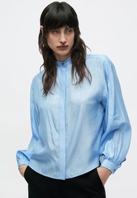 Svetlomodrá košeľa na gombíky s jemným pruhovaným vzorom, s vysokým golierom a nadýchanými rukávmi, vyrobená z hladkej, ľahkej látky.