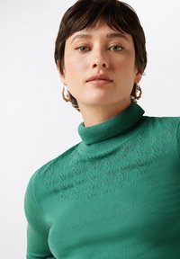 Groene coltrui met textuur van kantpatronen op de schouders, gemaakt van zachte stof. Kenmerkt zich door een aansluitende pasvorm en geribbelde nek.