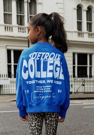 Blaues übergroßes Sweatshirt mit weißem "DETROIT COLLEGE" Schriftzug; verfügt über gerippten Kragen und Bündchen, kombiniert mit Leggings im Leopardenmuster.