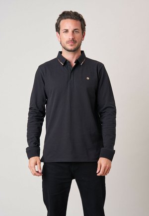 Man met kort krullend haar en baard draagt een zwart poloshirt met lange mouwen, staat voor een effen lichte achtergrond, kijkt recht vooruit met een neutrale gezichtsuitdrukking.