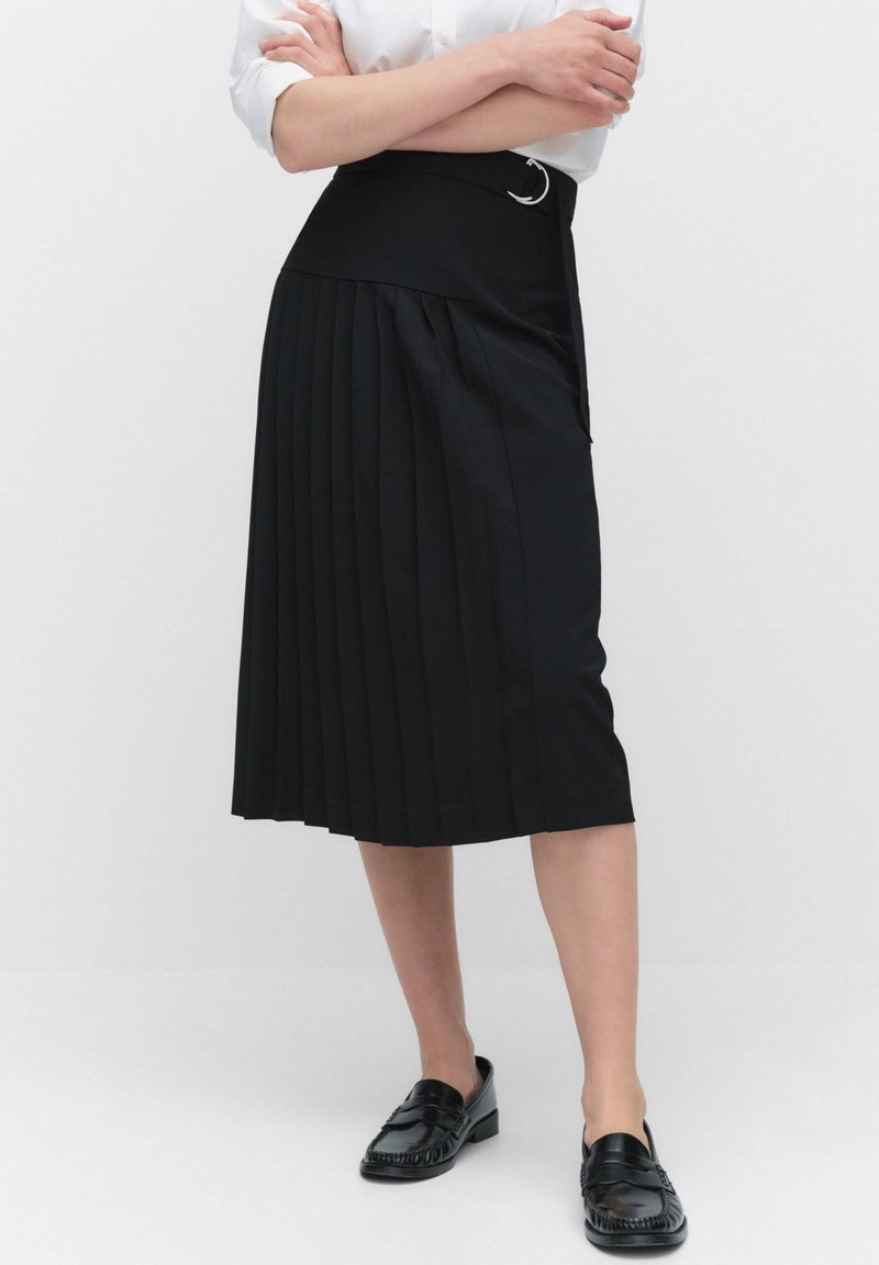 Vrouw in een zwarte plisé midi-skirt met een riem, een witte opgerolde blouse en zwarte loafers, staand met gekruiste armen.