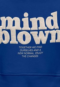 Blå hets med fet, upphöjd vit text som säger "mind blown" ovanför mindre vit text. Slät tyg med avslappnad passform och ribbat slut.