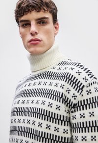 Strikket høyhalset genser i kremfarge med svarte geometriske mønstre. Har ribbestrikket hals og teksturert stoff med varierte horisontale striper.