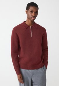 Pull bordeaux en mélange de laine avec un col montant zippé, des poignets et un ourlet côtelés, porté sur un pantalon gris, mettant en valeur une texture douce.