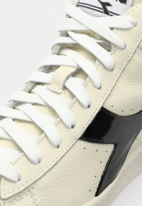 Beige leather sneaker with white laces, featuring a black glossy accent on the side and textured surface detailing.