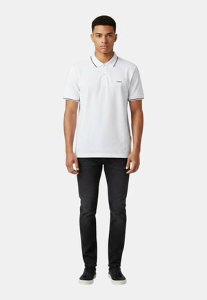 Giovane uomo in piedi, indossa una polo bianca a maniche corte con bordi neri, pantaloni neri slim fit e sneakers nere con suola bianca.