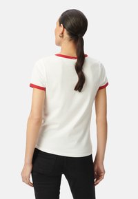 Camiseta de algodón blanca con un cuello y puños de manga en ribete rojo, diseño ajustado y espalda lisa. Combinada con jeans negros.