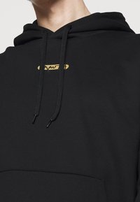Sudadera con capucha negra con bolsillo delantero, cordones negros y un pequeño logo dorado en el pecho. Tejido suave y liso con un ajuste relajado.