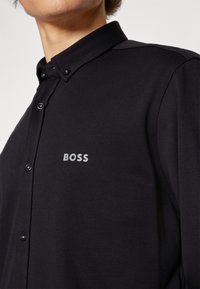 Schwarzes Baumwoll-Hemd mit Knopfleiste, strukturiertem Kragen, kontrastierenden Knöpfen und einem grauen "BOSS"-Logo auf der linken Brust.