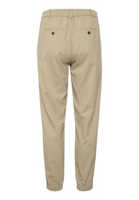 Beiga joggerbyxor med elastisk midja och muddar, med två bakfickor med svarta knappar. Slitstarkt, lättviktigt tyg.