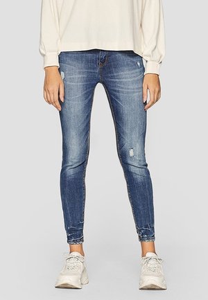 Persoon die vervaagde blauwe skinny jeans draagt met lichte slijtage, gecombineerd met witte sneakers en een lichtbeige top met lange mouwen.