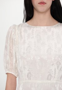 Bjela haljina s teksturiranim cvjetnim uzorkom, napuhanim rukavima i zaobljenim izrezom. Lagana i prozirna tkanina s delikatnim detaljima.