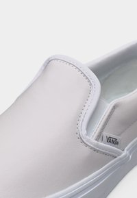 Λευκό slip-on παπούτσι Vans με λεία επιφάνεια, ελαστικές πλαϊνές λεπτομέρειες και μικρή ετικέτα Vans στο πλάι κοντά στο άνοιγμα.