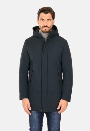 Cappotto nero con cappuccio, chiusura con zip e bottoni a pressione, due tasche laterali e una texture liscia. Indossato sopra un dolcevita grigio e jeans.