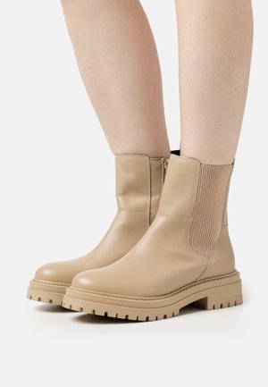 Stivaletti in pelle beige sopra la caviglia con pannelli laterali rigati, suola spessa e finitura liscia. Il design presenta hardware minimale e una silhouette snella.