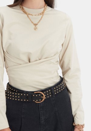 Beige top met lange mouwen en overslagdetail, zwarte gesp met studs en gouden gesp op zwarte broek, laagjes gouden kettinkjes en een gouden armband.