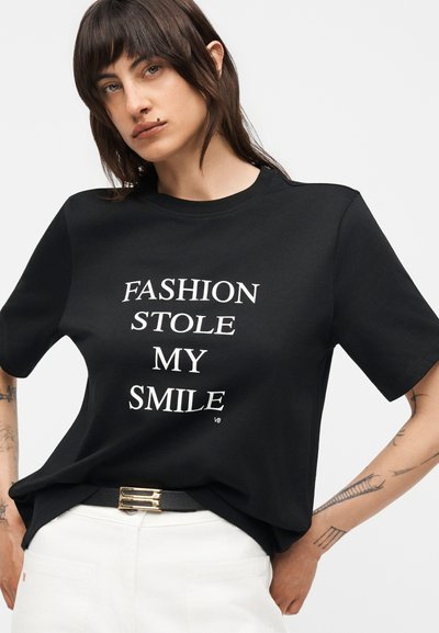 Άτομο με μαύρο μπλουζάκι που γράφει λευκά "FASHION STOLE MY SMILE", φορεμένο με λευκό παντελόνι και μαύρη ζώνη με χρυσή αγκράφα.