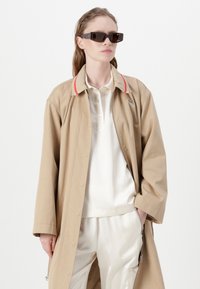 Beige ulkoilutakki punaisilla yksityiskohdilla, nappikiinnitys. Valkoinen polo paidan alla ja kreeminväriset satiinisedanssit. Tummat suorakaiteen muotoiset aurinkolasit viimeistelevät ilmeen.