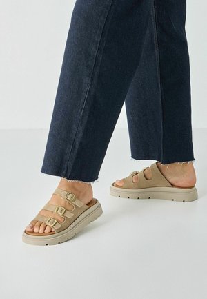 Beżowe sandały ze skóry zamszowej posiadają trzy regulowane paski z pozłacanymi sprzączkami, masywną, kremowo-białą podeszwę oraz teksturowaną wkładkę dla wygody.