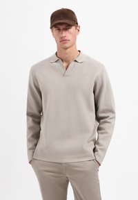 Lichtbeige poloshirt met lange mouwen, V-hals en een zachte textuur. Gecombineerd met beige broek en een bruine pet. Eenvoudig en modern ontwerp.