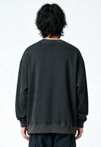 Personne aux cheveux noirs ébouriffés portant un sweat-shirt noir délavé surdimensionné et un pantalon noir, vue de dos sur un fond blanc uni.