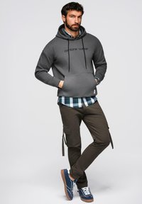 Mężczyzna w szarej bluzie z kapturem z napisem "ombre wear", niebieskiej koszuli w kratę, brązowych spodniach cargo i niebieskich trampkach, stojący z rękami w kieszeniach.