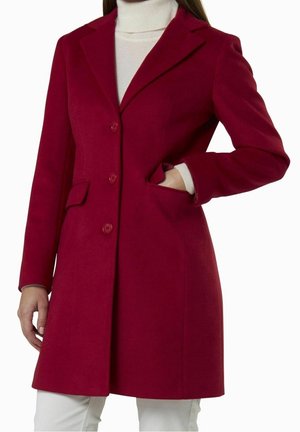 Femme portant un manteau rouge boutonné avec un col, une main dans la poche, par-dessus un col roulé blanc et un pantalon blanc.