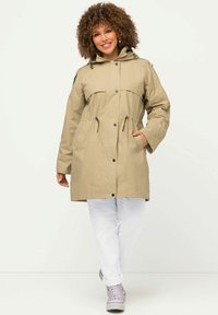Khaki parka takki, joka sisältää kiristettävän vyötärön, suuret etutaskut, nappisulkemisen ja huppua. Tyylitelty valkoisten housujen ja harmaiden kenkien kanssa.