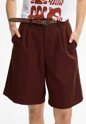 Persona que lleva pantalones cortos de tiro alto de color rojo oscuro con bolsillos laterales y un cinturón tejido marrón, combinado con una camiseta blanca con estampado gráfico.