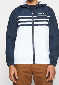 Veste coupe-vent bleu marine et blanche avec des rayures horizontales, fermeture éclair à l'avant, capuche ajustable et poignets élastiques, associée à un pantalon marron.