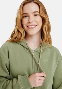 Grüne Zip-Hoodie mit Kordelzug. Weicher Stoff, gerippte Bündchen und ein kleines Logo auf der Brust. Das Modell hat lockiges Haar und hält den Kordelzug.