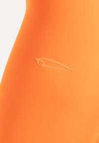 Collants de sport orange en tissu extensible, avec un logo en relief. La surface présente une texture lisse et mate.