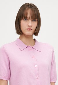 Polo shirt rosa chiaro a maniche corte con colletto, caratterizzato da una texture lucida e sei bottoni rosa lungo la parte anteriore. Design semplice e strutturato.