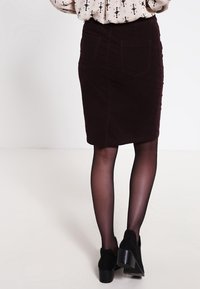 Burgunder Kordrock im Bleistiftstil mit hinteren Taschen, strukturiertem Oberflächen und taillierter Passform. Getragen mit schwarzen Stiefeletten und transparenter Strumpfhose.