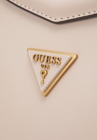 Beige læderoverflade med et trekantet metal Guess USA-logo i guld- og cremefarver, der har et spørgsmålstegn.