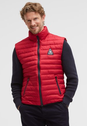 Smiling man with curly hair wears a red quilted sleeveless vest over a dark long-sleeve sweater, hands in vest pockets, light background.