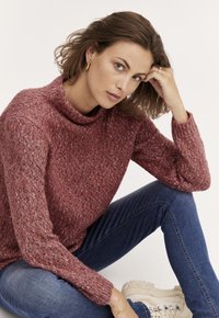 Adīts džemperis dziļā rozā krāsā ar augstu apkakli; tekstūra ar krāsu traipiem. Saskaņots ar pieguļošām zilu džinsām un gaišiem masīviem sporta apaviem.