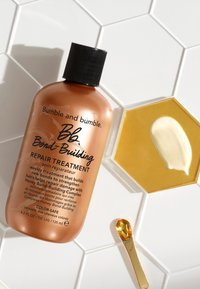 Brun plastflaska med svart kapsyl märkt "Bumble and bumble Bb. Bond-Building Repair Treatment," bredvid en gyllene hexagonal skål med kräm.