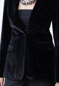 Kvinde iført en sort fløjlsblazer over en sort tube top, accessoriseret med en chunky metallic sølv perlehalskæde.