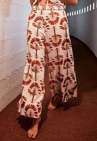 Pantalon à jambes larges en tissu crème avec un motif floral rouge et marron. Il présente une taille à cordon et une texture douce.