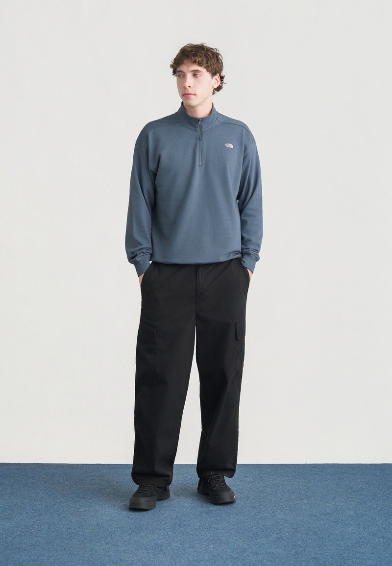 Blue zip-up fleece pullover, black cargo pants with pockets, shown on a person standing on a blue carpet with a plain light background.