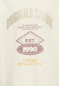 Kremowy materiał z nadrukowanym tekstem: "ORIGINALS STUDIO," "EST. 1990," oraz "LOCAL + COLLECTIVE" w naturalnych odcieniach.