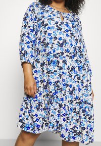 Bloemendress in blauwtinten met een gelaagd ontwerp, keyhole-halslijn en driekwartmouwen van lichte stof.