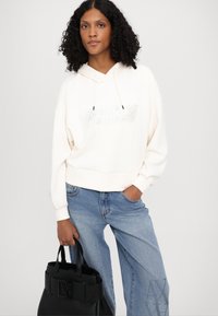 Felpa crema con cappuccio, dal taglio rilassato e con accento del logo, abbinata a jeans in denim azzurro chiaro. Borsa nera dal design strutturato.