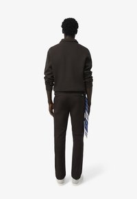 Donkerbruine trui met een kraag, gecombineerd met bijpassende broek. Een blauw-wit gestreepte sjaal is om de taille geknoopt. Witte sneakers zijn zichtbaar.