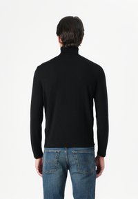 Μαύρο πουλόβερ με μακριά μανίκια και ψηλό γιακά, φτιαγμένο από μαλακό πλεκτό ύφασμα, με εφαρμοστή γραμμή και λεία υφή. Φοριέται με μπλε τζιν.
